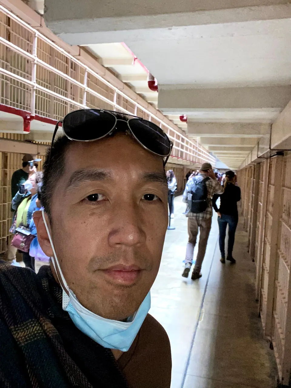 A close selfie of a man with sunglasses on his head and a mask pulled down, with visitors walking away in a cell-block corridor behind him.