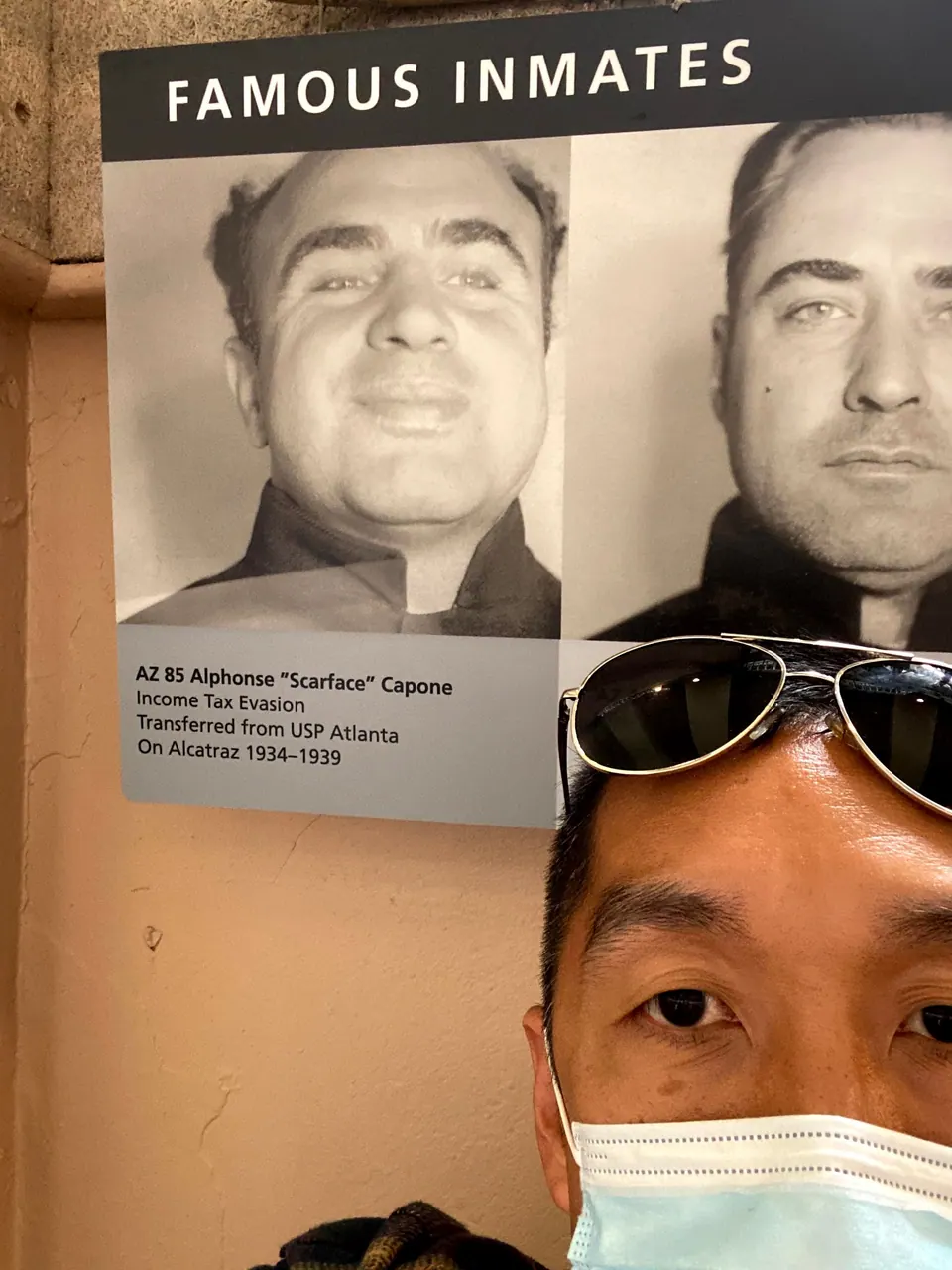 Half of a man's face with sunglasses pushed up and a mask beside a Famous Inmates panel with mugshots and a caption naming Al Capone.