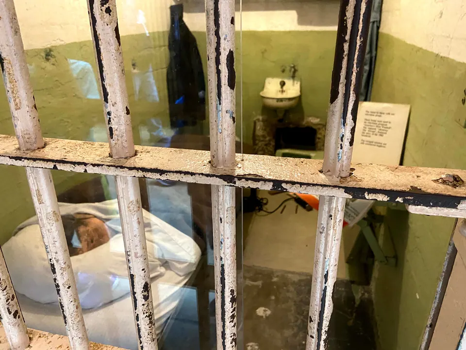Through chipped, peeling cell bars, a green-walled cell with a dummy head on the pillow behind glass, a sink and a small card.