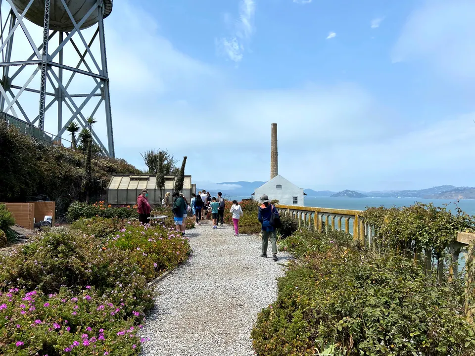 A gravel path through flowering gardens toward a tall chimney, with a steel water tower at left, visitors ahead and hills across the bay.