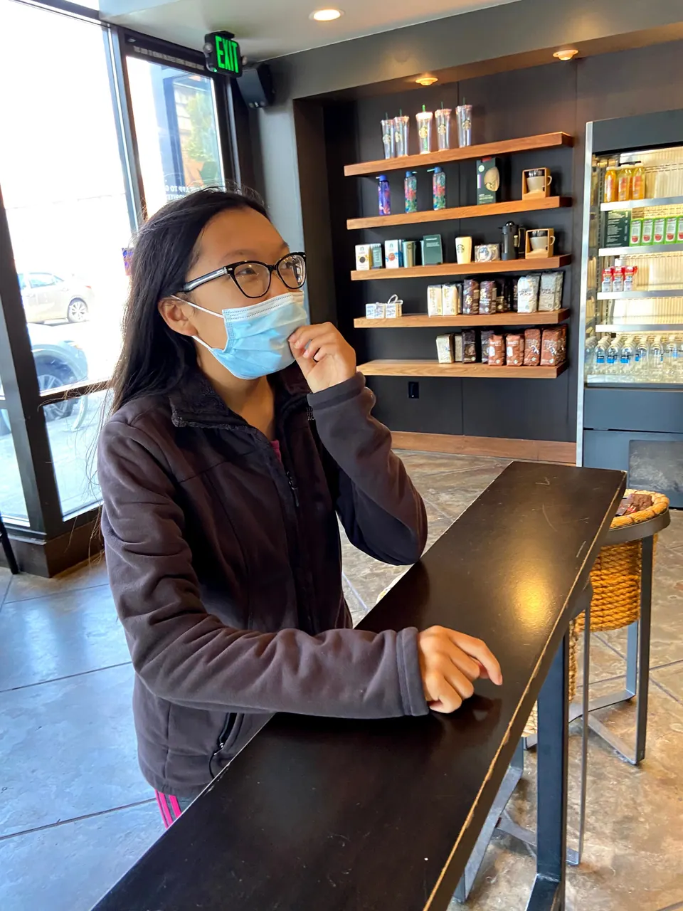 A masked girl stands at a black counter in a coffee shop with shelves of tumblers and packaged coffee behind her and sunlit windows at left.