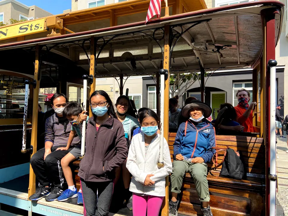 Passengers sit and stand on the open side of a cable car, two masked girls at the front and a woman in a wide-brimmed hat on the bench.