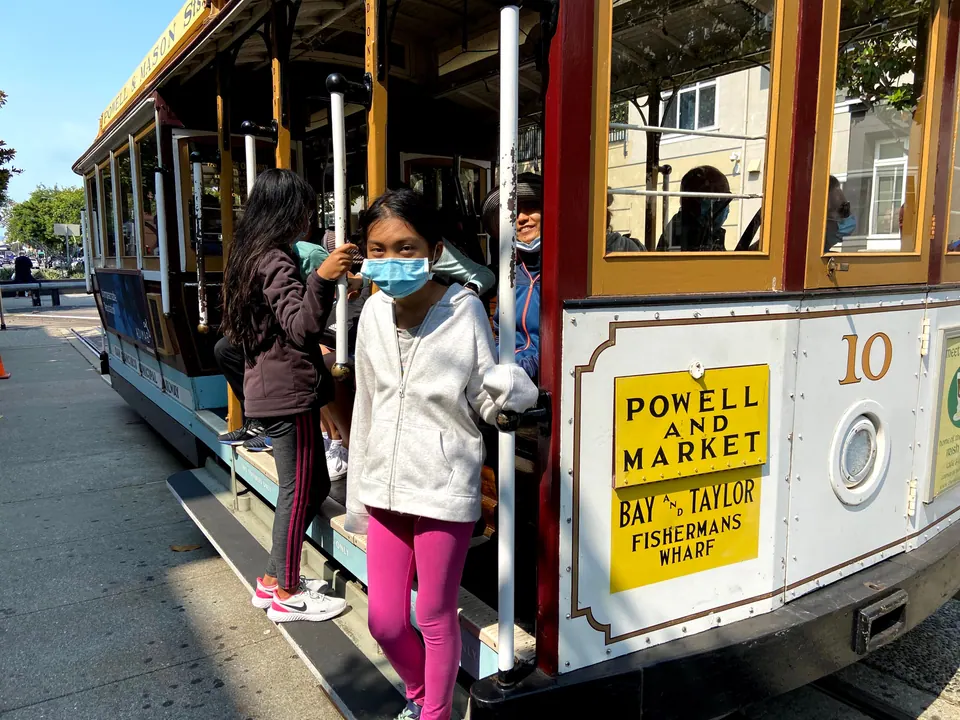 A masked girl in a white hoodie holds the pole at the step of cable car number 10, with its yellow Powell and Market, Bay and Taylor sign and another girl behind her.