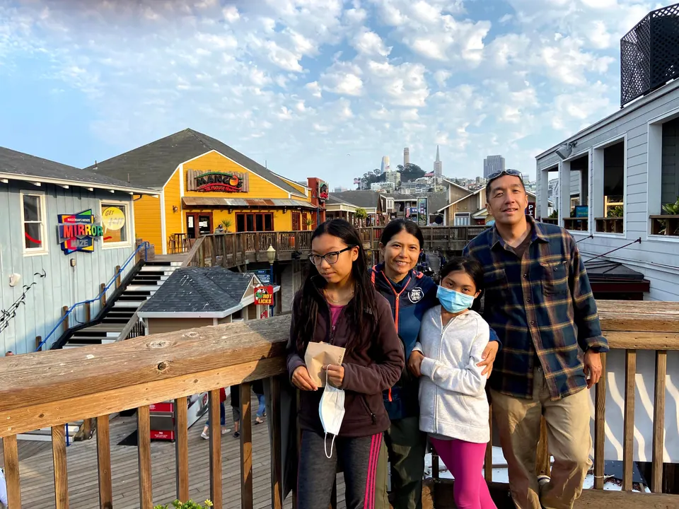 Four people pose at a wooden railing on a pier in soft evening light, with a mirror maze sign, a yellow building and a tower on the hill behind.