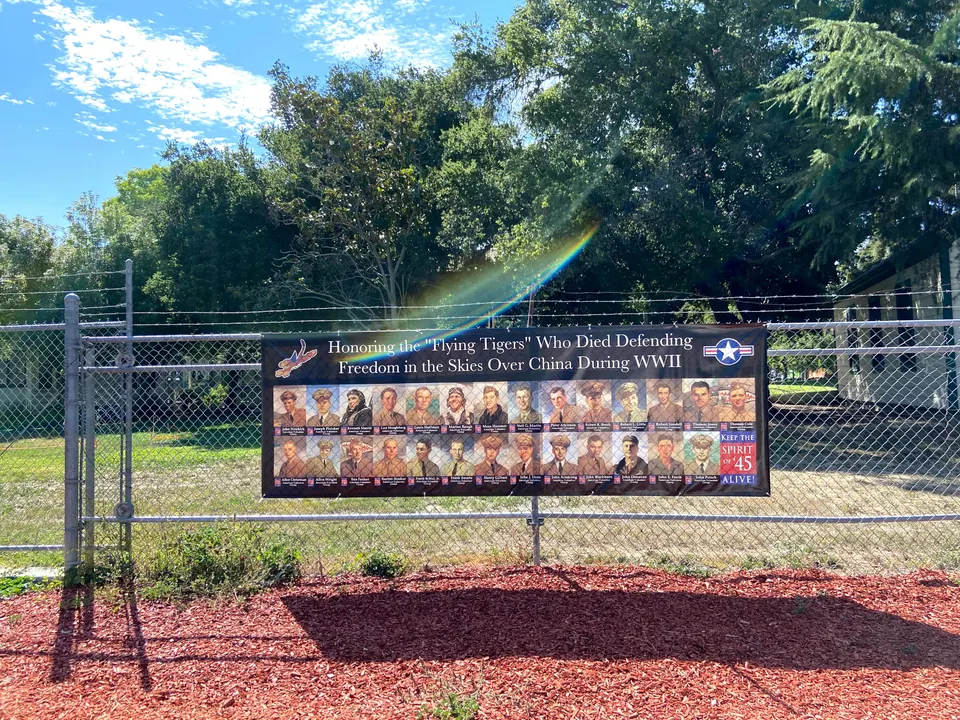 A banner on a chain-link fence honoring the Flying Tigers who died defending China, with a rainbow lens flare, a lawn and tall trees behind.