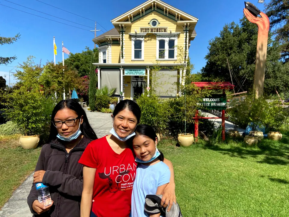 A woman and two girls stand on a lawn before a yellow Victorian building marked Viet Museum, with flags and a red garden gate beside them.