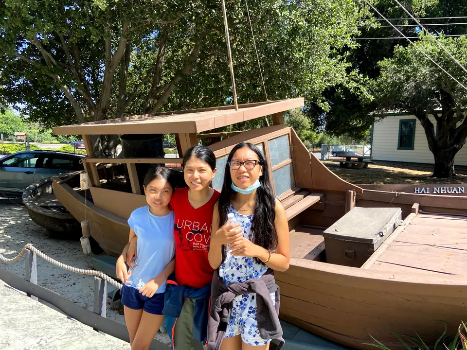 A woman and two girls pose beside a large wooden boat with the name Hai Nhuan painted on its side, under big shade trees.