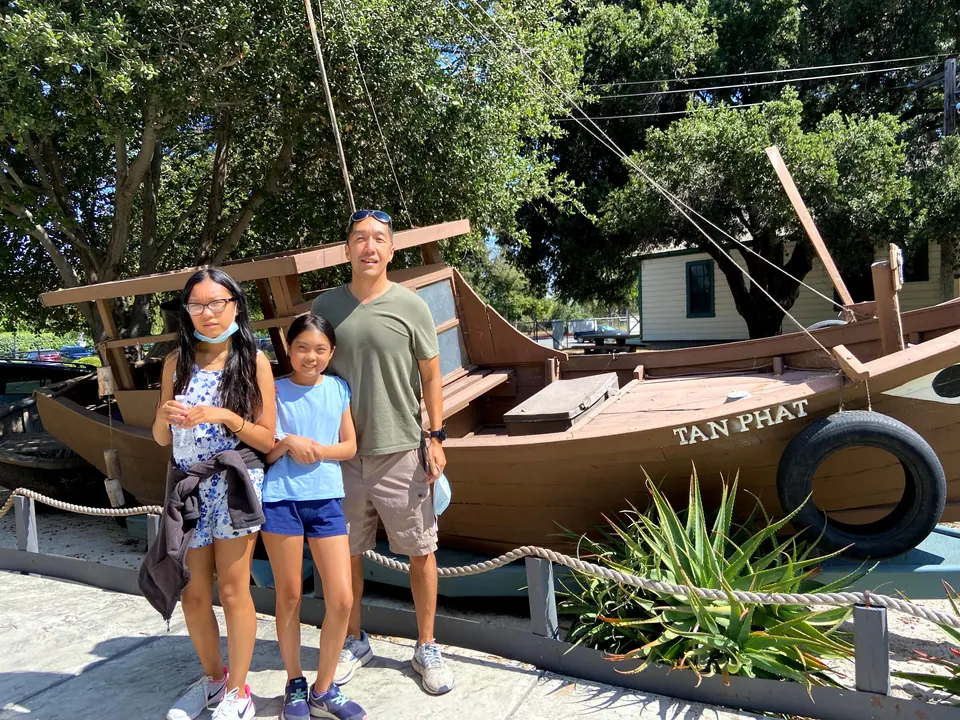 A man and two girls stand in front of a wooden boat named Tan Phat, with a tire fender on its side and an aloe plant at its foot.