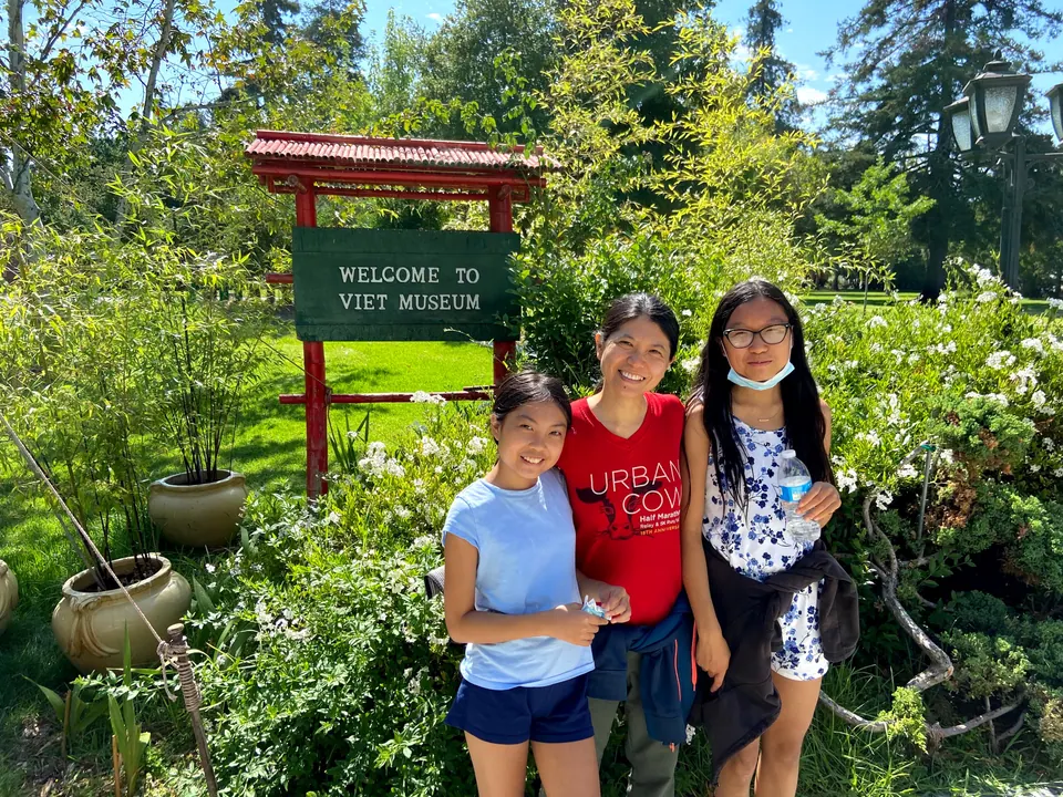 A woman and two girls smile in front of a green Welcome to Viet Museum sign under a red roof, among white flowering shrubs.