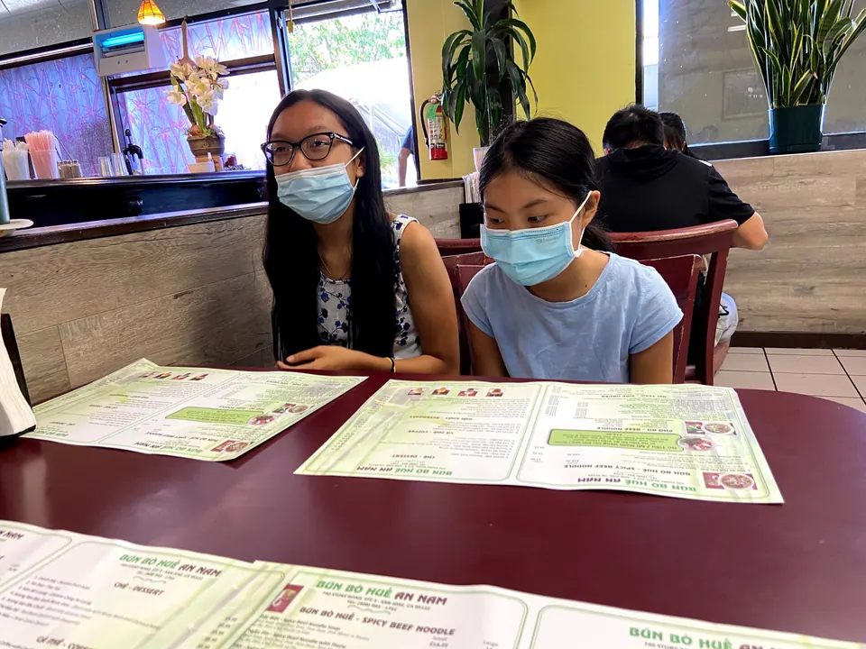 Two masked girls sit at a maroon table with green laminated menus in a small restaurant with yellow walls and big potted plants.