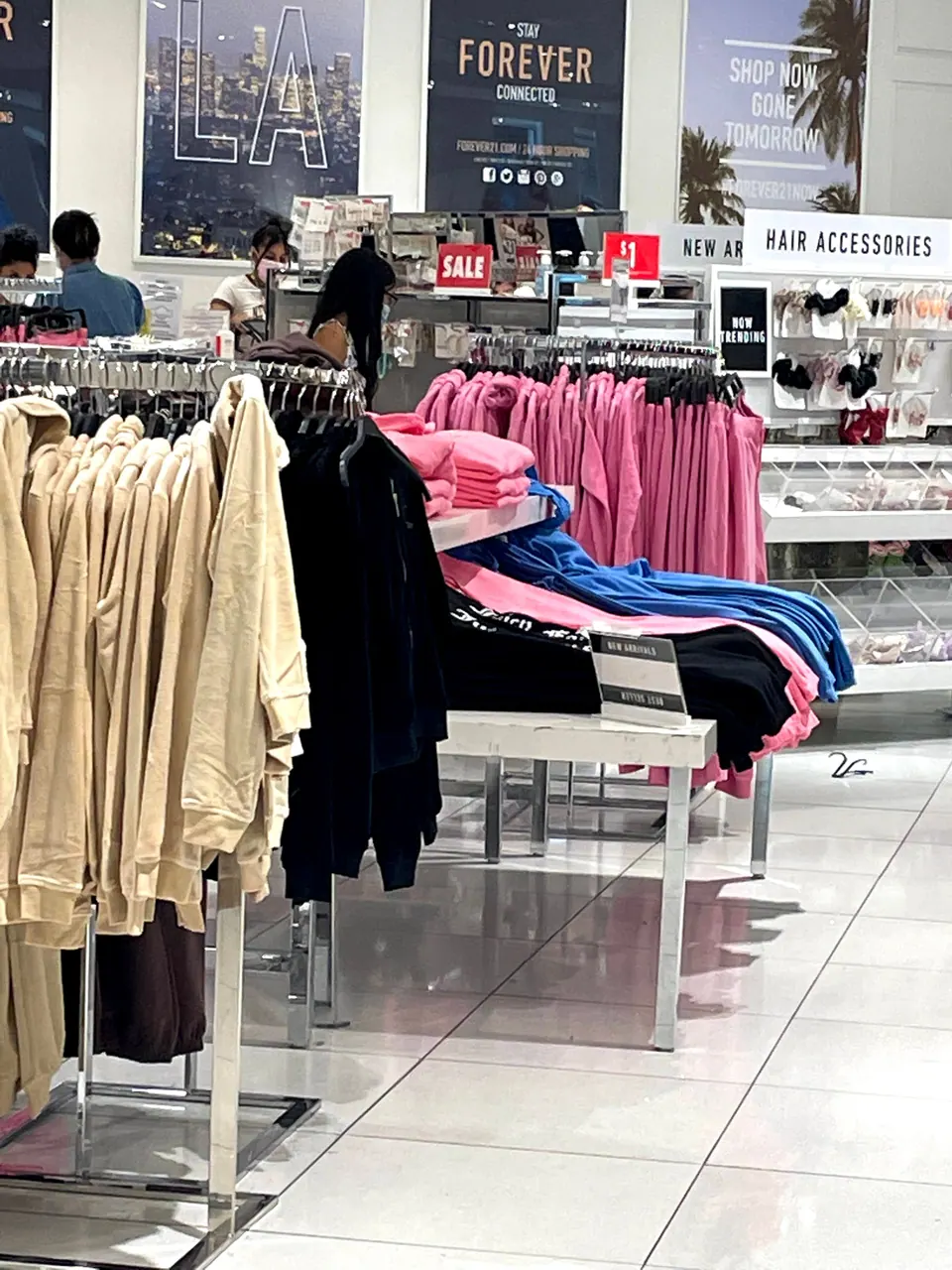 Racks of beige and black sweatshirts and a table of pink and blue tops inside a clothing store, with a Sale sign and posters behind.