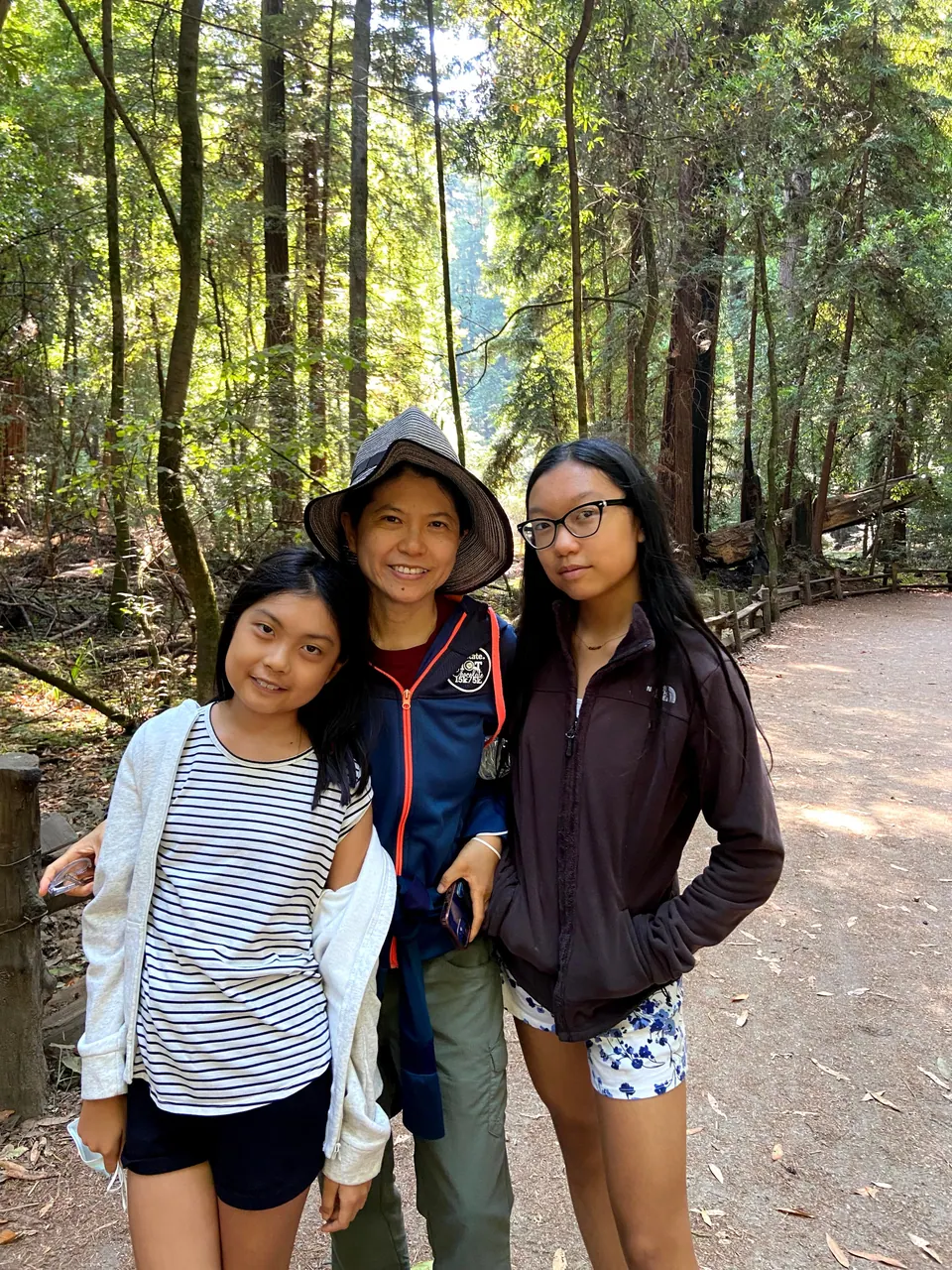 A woman in a wide-brimmed hat stands between two girls on a dirt trail lined with tall trees and a rustic fence.