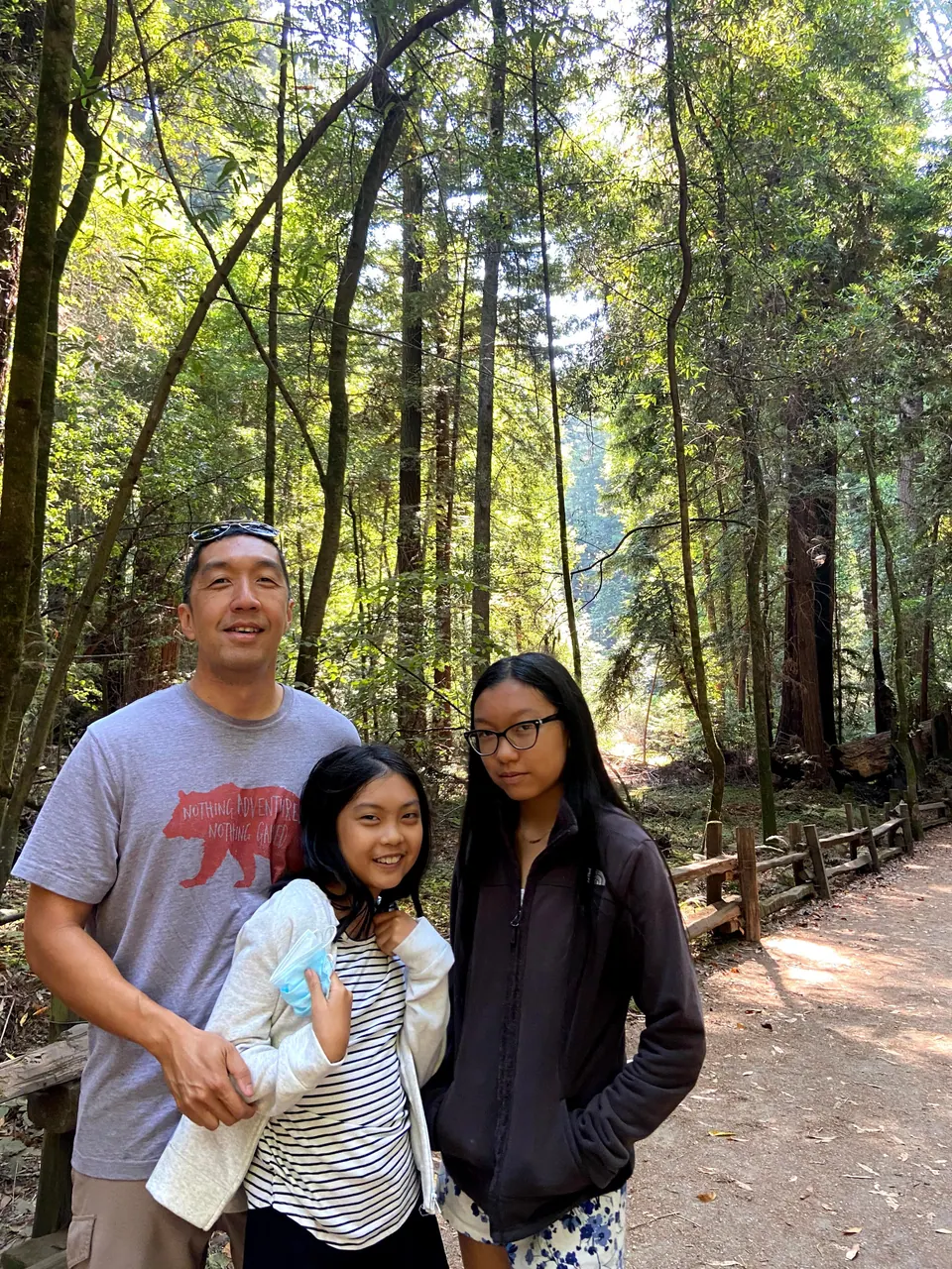 A man with sunglasses on his head and two girls pose on a trail with tall redwood trunks and a split-rail fence behind them.