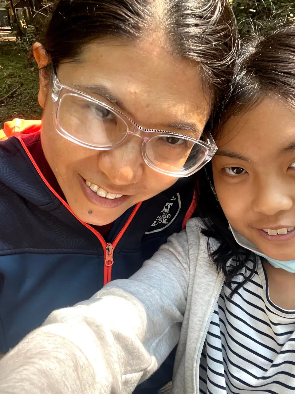 A close selfie of a woman in clear-framed glasses and a navy jacket cheek to cheek with a smiling girl in a striped top, her mask under her chin.