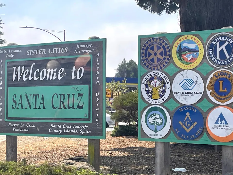 A green Welcome to Santa Cruz sign listing its partner cities beside a board of service-club emblems including Rotary, Kiwanis and Lions.