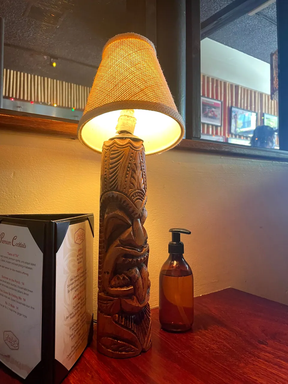 A carved wooden tiki lamp with a burlap shade beside a cocktail menu card and a hand-soap bottle on a dark wood table, with bamboo walls in the window behind.