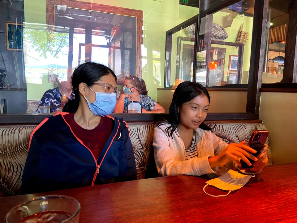 A woman in a mask and a girl looking at her phone sit side by side in a palm-print booth, with a drink glass in the foreground.