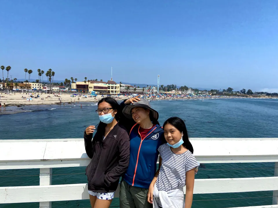 A woman in a wide-brimmed hat and two masked girls pose at a white wharf railing, with a sandy beach, the Boardwalk buildings and a roller coaster across blue water behind them.