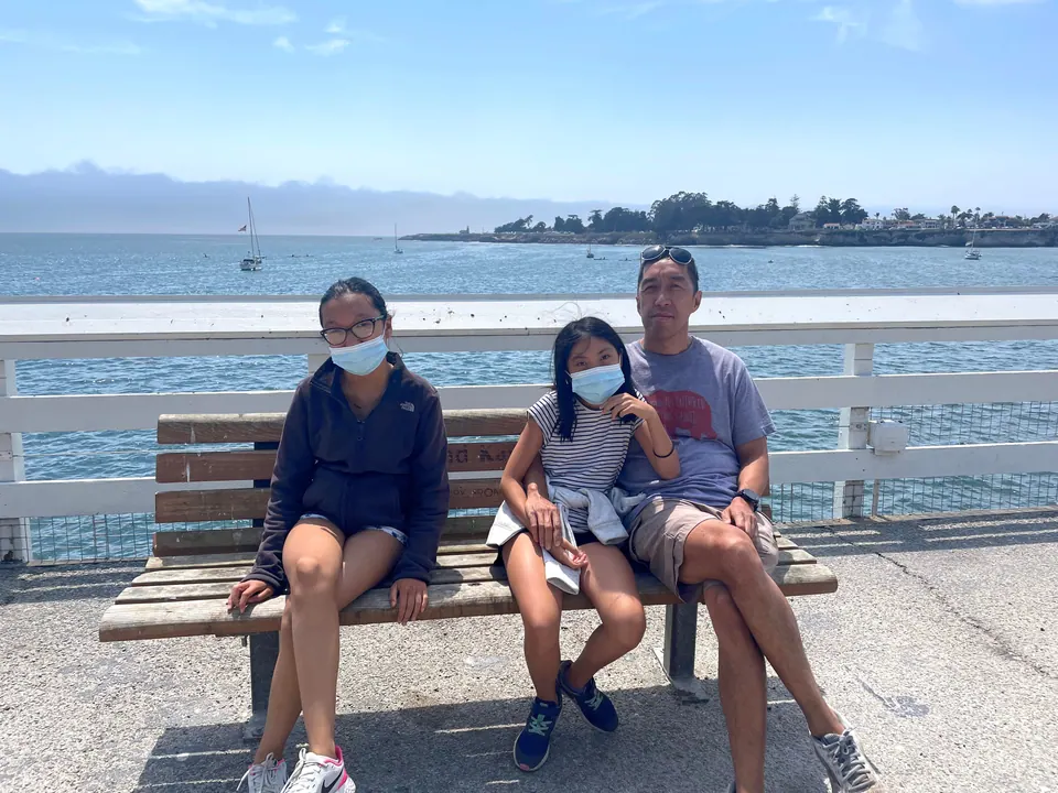 Two masked girls and a man with sunglasses on his head sit close together on a wooden bench at a white railing, with sailboats and coastal cliffs behind.