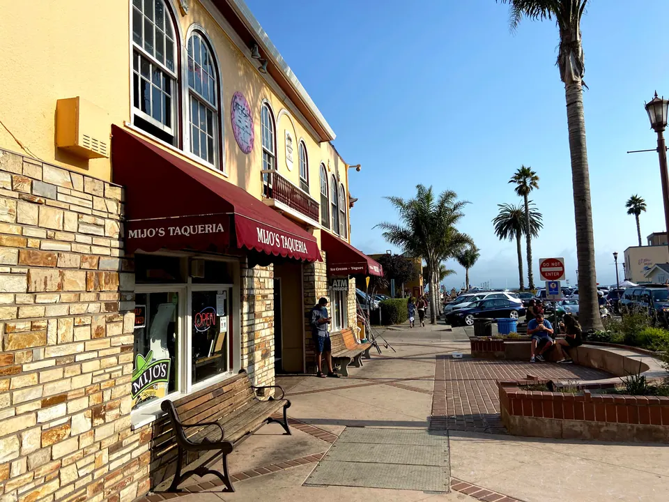 A cream and stone corner building with a maroon Mijo's Taqueria awning and an Open sign, with a bench outside, palm trees and a village square in evening sun.