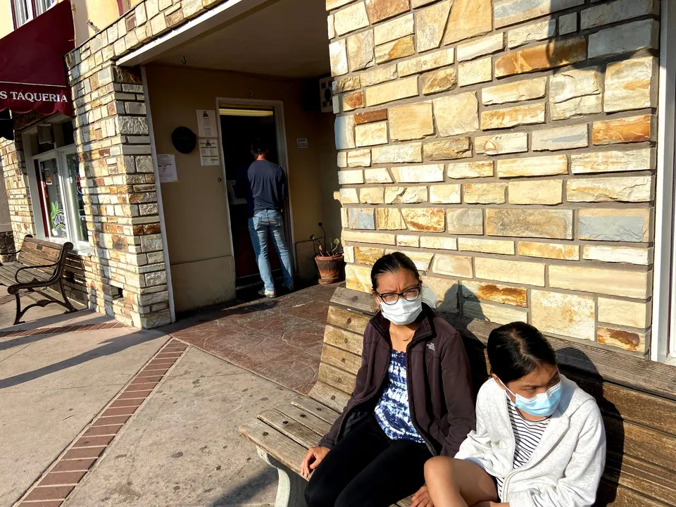 Two masked girls sit on a wooden bench against a stone wall outside a taqueria while a man in a navy shirt steps in through the door behind them.