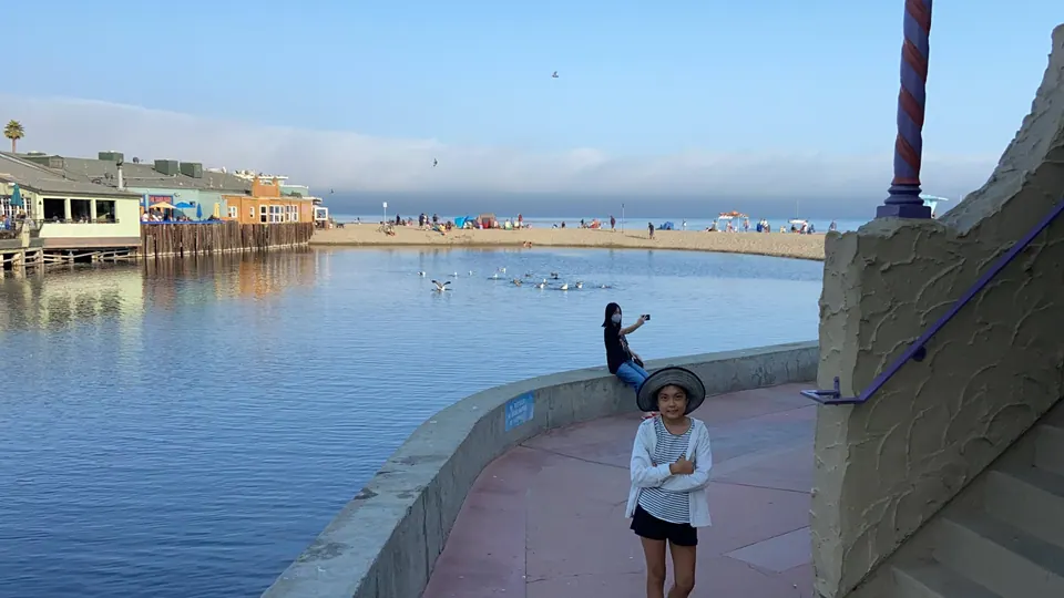 A girl in a sun hat stands with her arms crossed on a pink walkway beside a sea wall, with another girl behind her photographing the lagoon and beach.