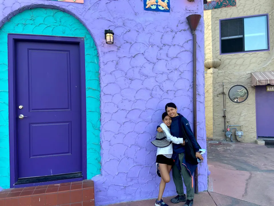 A woman and a girl hug against a lavender scalloped stucco wall with a purple door framed in turquoise, the girl's sun hat in her hand.