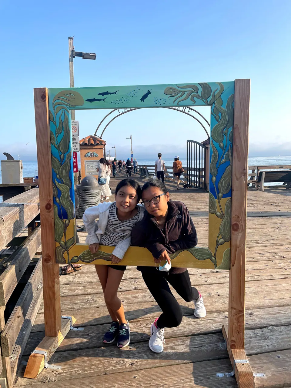 Two girls lean on the bottom rail of a hand-painted wooden frame decorated with kelp, fish and a turtle, posing on a wharf's boards with an arched gate and strollers behind them.