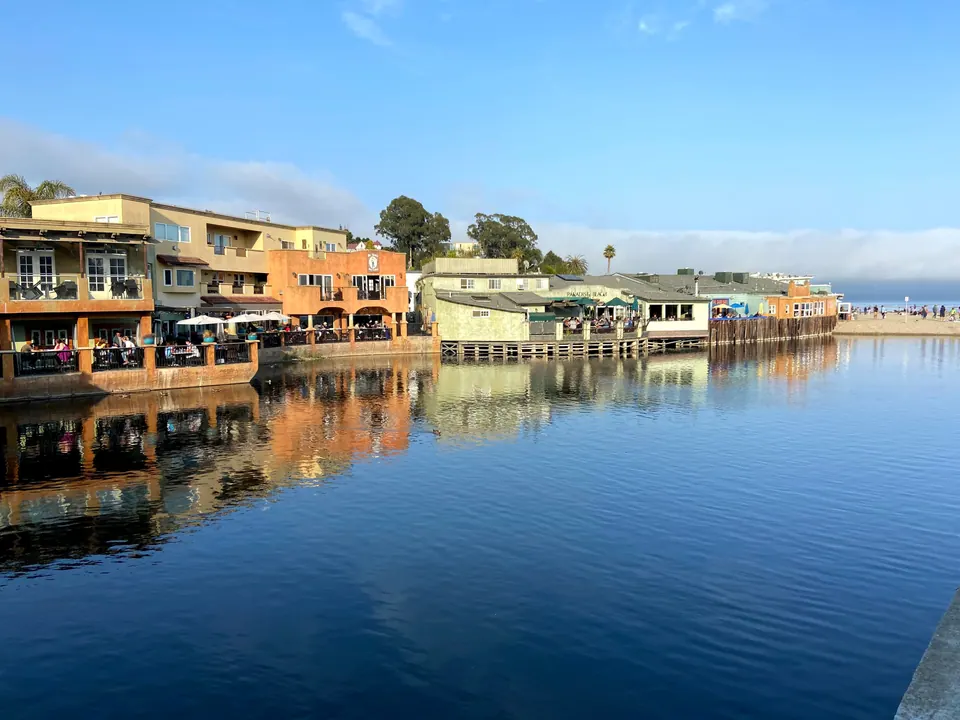 Terraced waterfront restaurants with diners on their decks are reflected in still water, with a fog bank behind the beach.