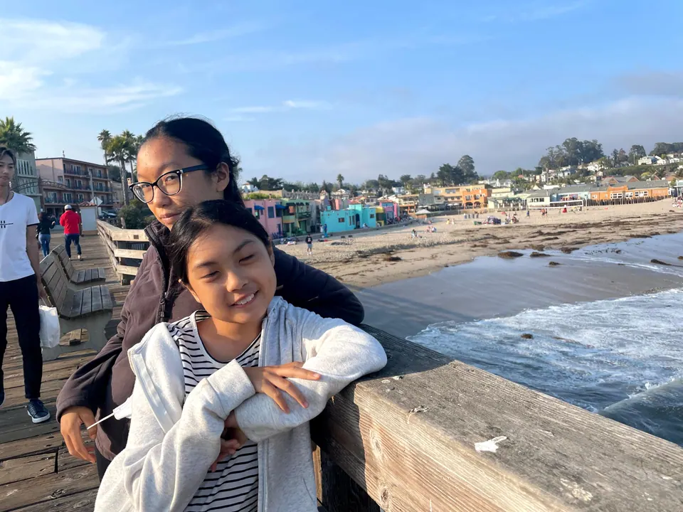 Two girls lean together on the wooden railing of a wharf in low sun, with the beach, surf and pastel cottages behind them.
