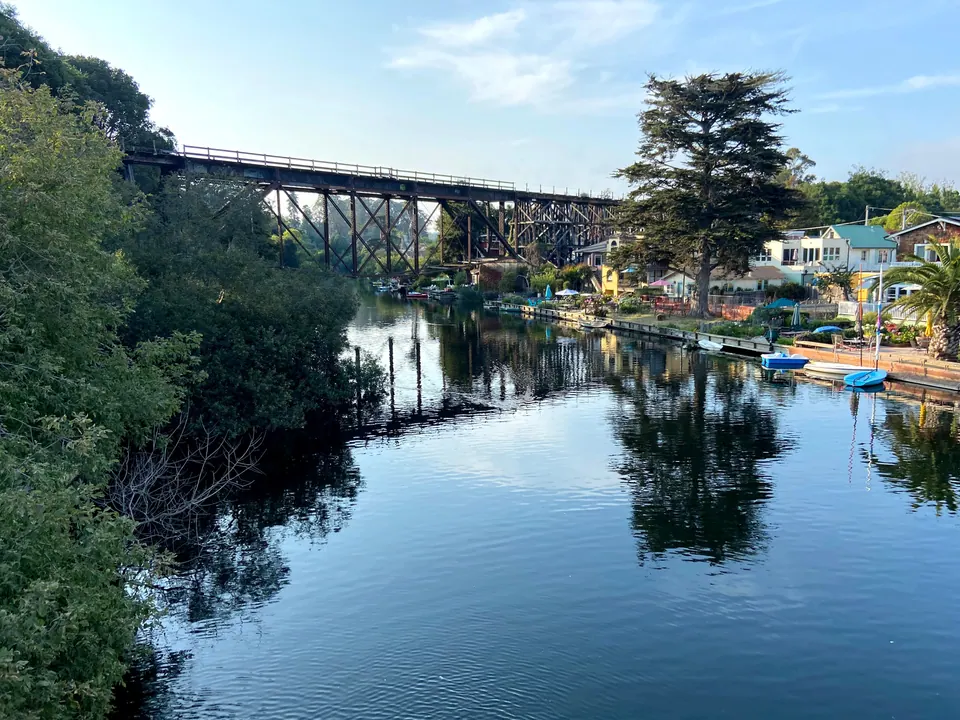 A dark iron railroad trestle crossing a creek above still, green-fringed water and creekside houses with small boats, in soft evening light.