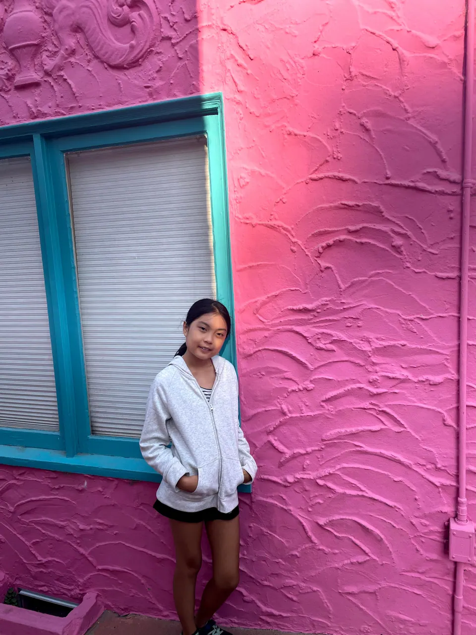 A girl in a white cardigan stands with her hands in her pockets against a bright pink stucco wall with a turquoise-framed window and a relief carving above.