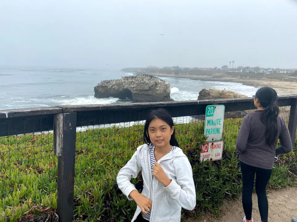 A girl in a white hoodie stands at a fence with an arch rock and cormorants behind her, another girl looking out to sea beside a 20 Minute Parking sign.