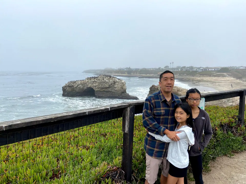 A man in a blue plaid flannel shirt hugs a girl in a white hoodie, a second girl in glasses beside them, at a railing above a beach with a rock arch in fog behind.