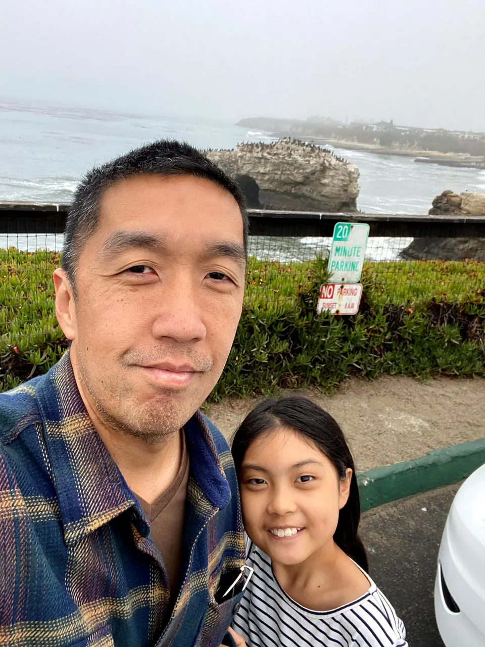 A selfie of a man in a blue plaid flannel shirt and a smiling girl in a striped top at an overlook, with an arch rock and a 20 Minute Parking sign behind them.