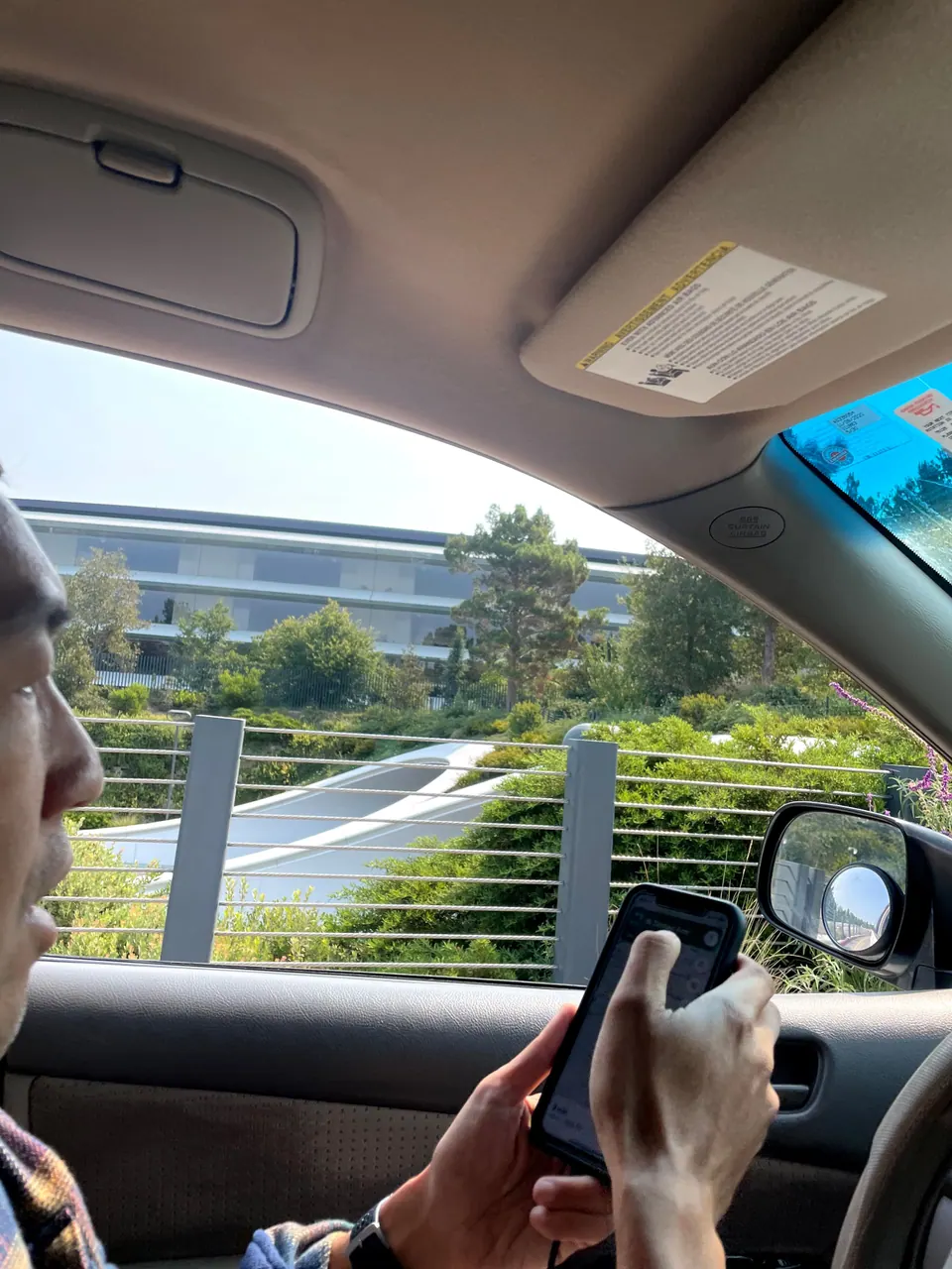 Through a car window, a glass ring building above a hillside of shrubs and a cable-rail fence, with a man's hands holding a phone in the foreground.