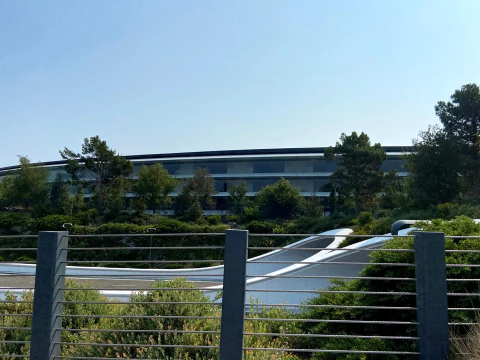 The curved glass ring of a large building seen over a cable-rail fence and landscaped shrubs, with a white service road curving in front, under a pale sky.