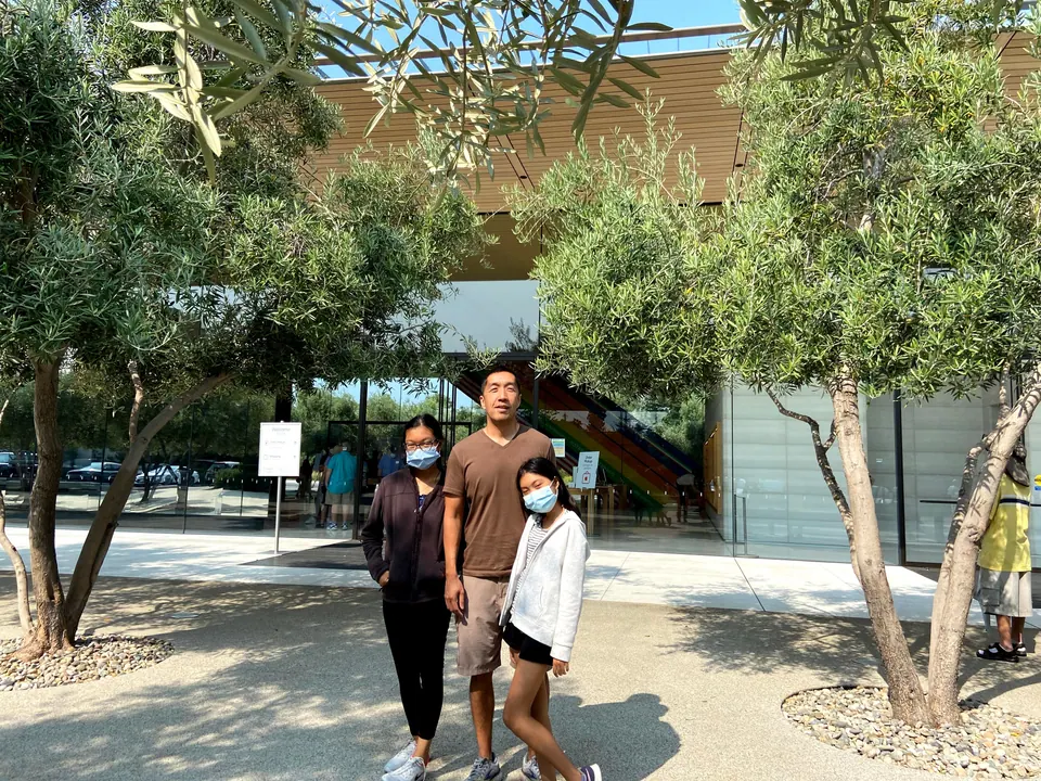A man in a brown T-shirt stands between two masked girls under olive trees in front of a glass pavilion, with a rainbow staircase glimpsed inside.