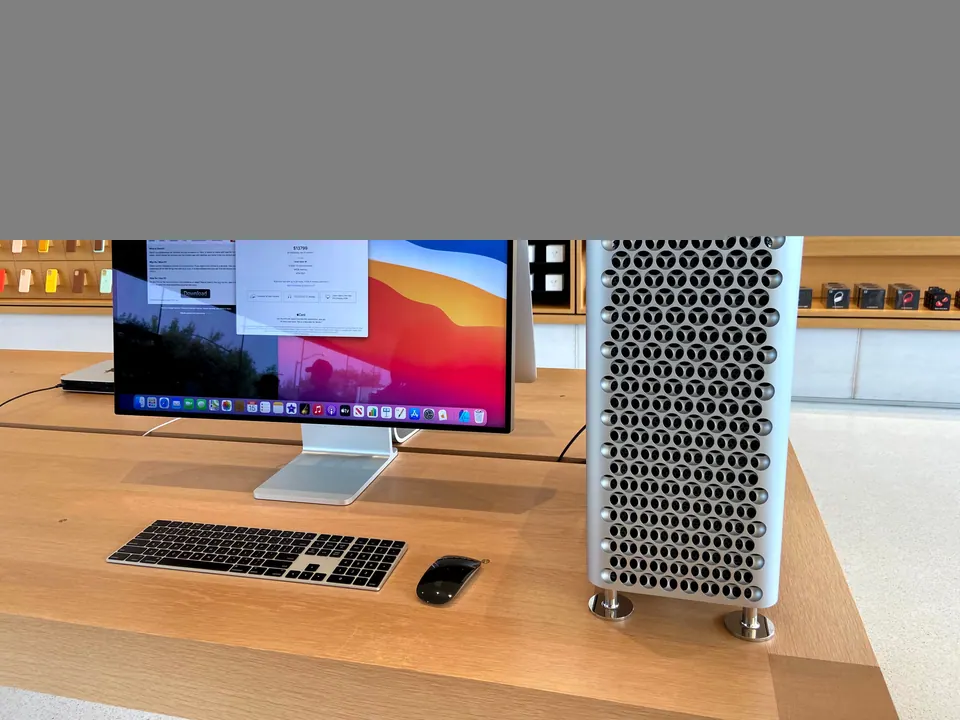 A Mac Pro tower with a lattice case beside a large display, a keyboard and a mouse on a pale wood table, with walls of phone cases and headphones behind.