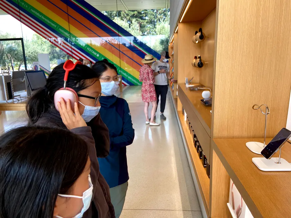 A girl holds pink over-ear headphones to her ear while a woman looks on beside her and another girl fills the foreground, with a rainbow staircase and display shelves behind.