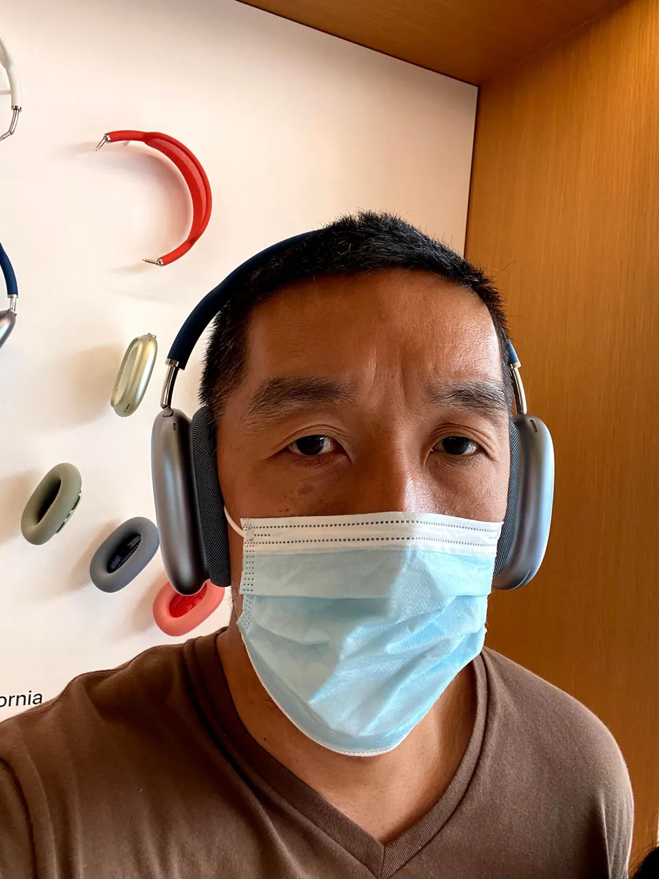 A selfie of a man in a face mask and brown T-shirt wearing silver over-ear headphones in front of a wall of colored ear cushions and headbands.