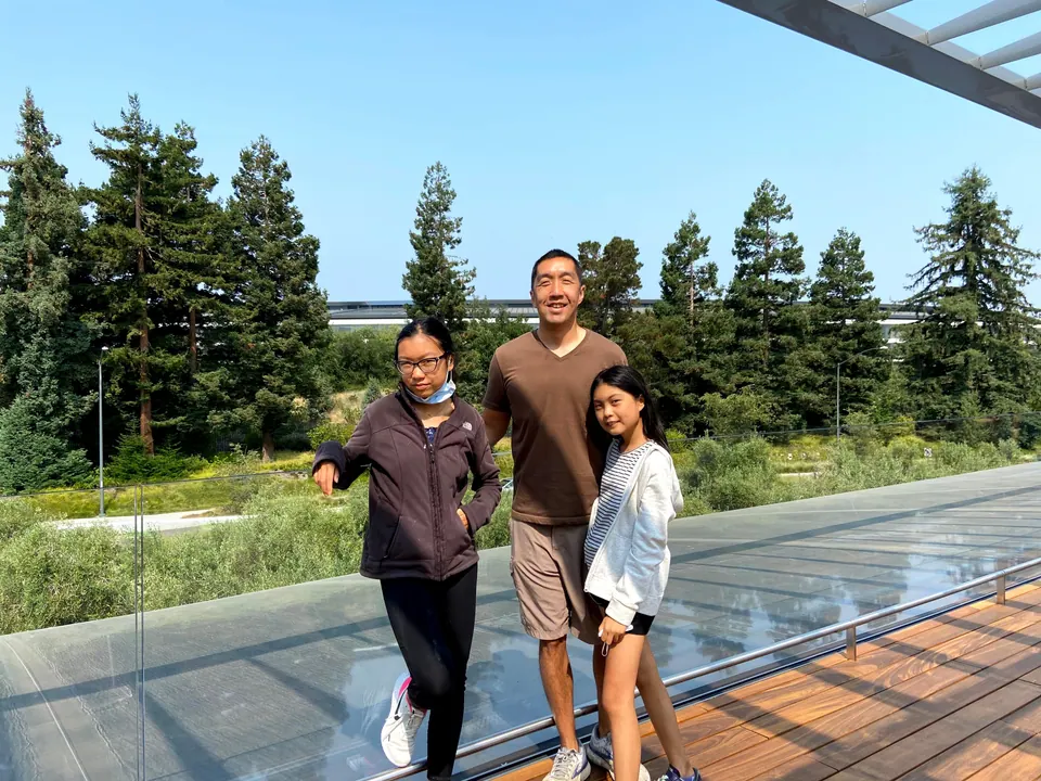 A man in a brown T-shirt and two girls stand on a wooden rooftop terrace beside a glass railing, with tall redwoods behind them and a long curved building in the distance.