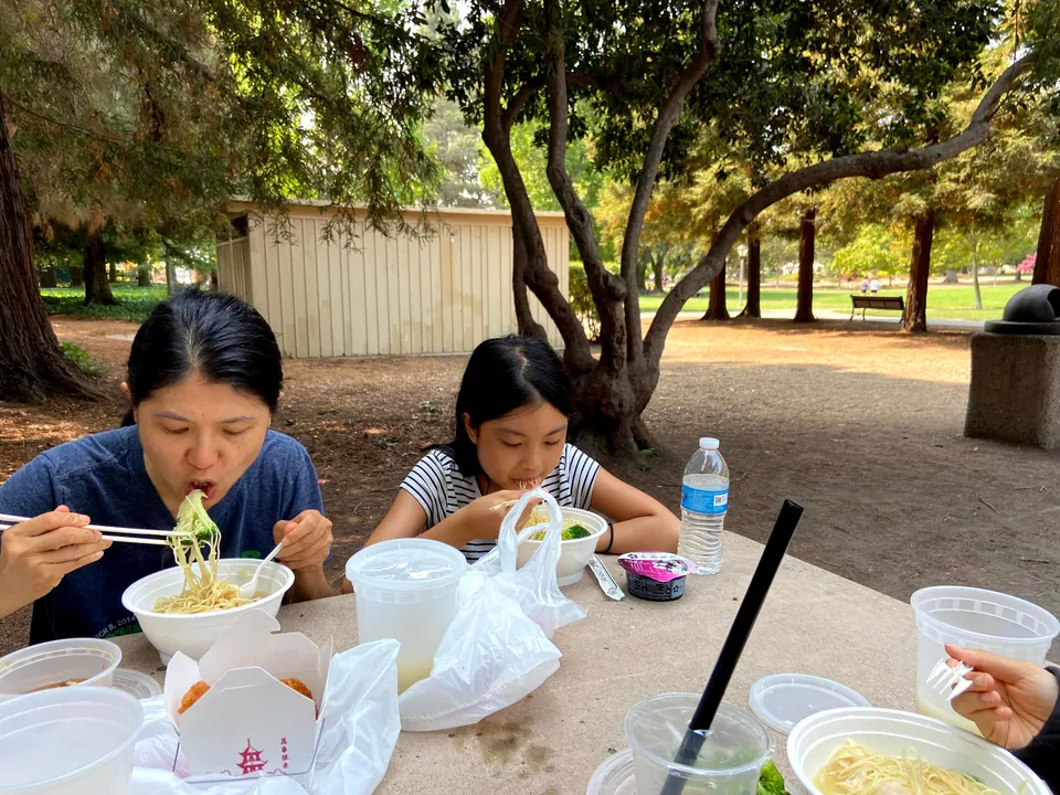A woman eats noodles with chopsticks beside a girl in a striped top at a stone picnic table under trees, with takeout bowls, cups, a water bottle and a takeout carton spread around.