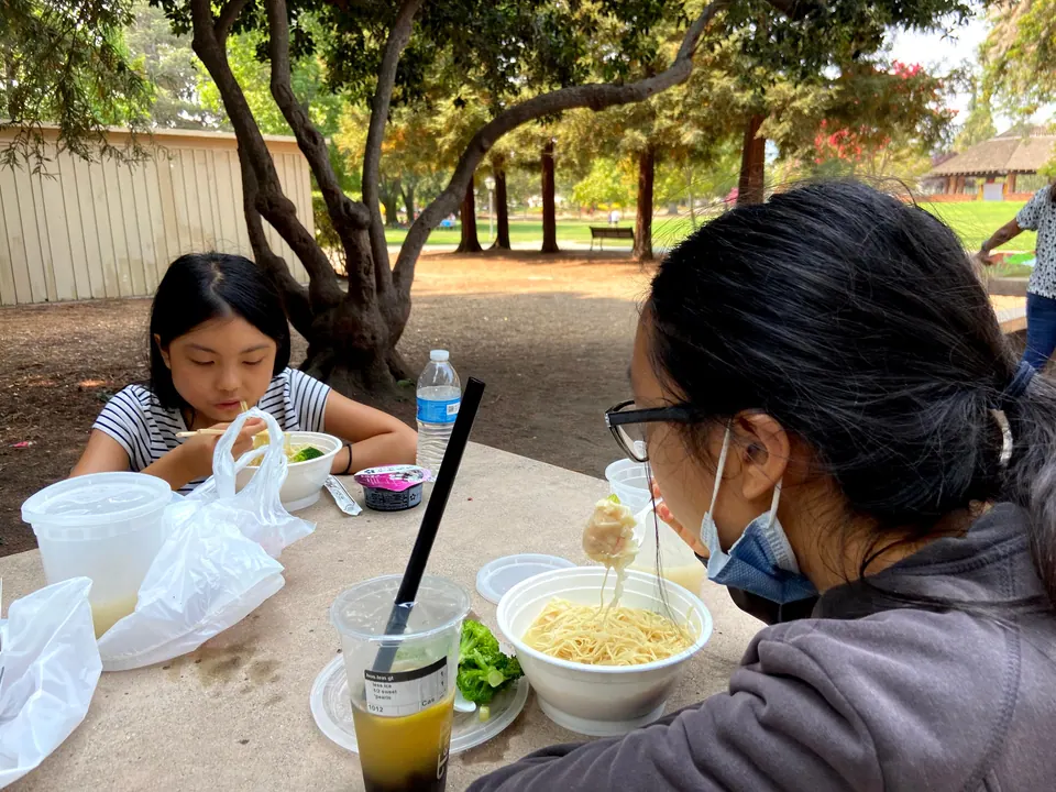 A girl in a striped top eats noodles across the table from a masked girl in glasses lifting a forkful of noodles, with a bubble-tea cup and a black straw between them and a park lawn behind.