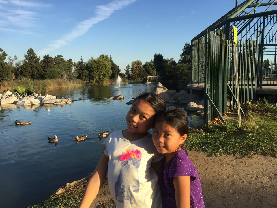 Two girls lean their heads together beside a pond, with ducks on the water and a fountain behind them.