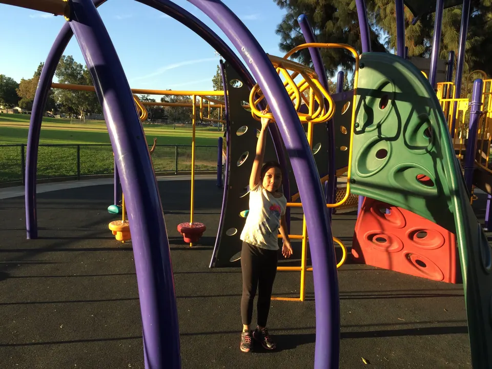 A girl in a white T-shirt and black leggings reaches up to a yellow bar inside the purple arches of a playground, a green lawn behind.