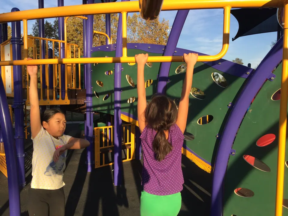 Two girls hang from the yellow bars of a purple and yellow playground climbing frame.