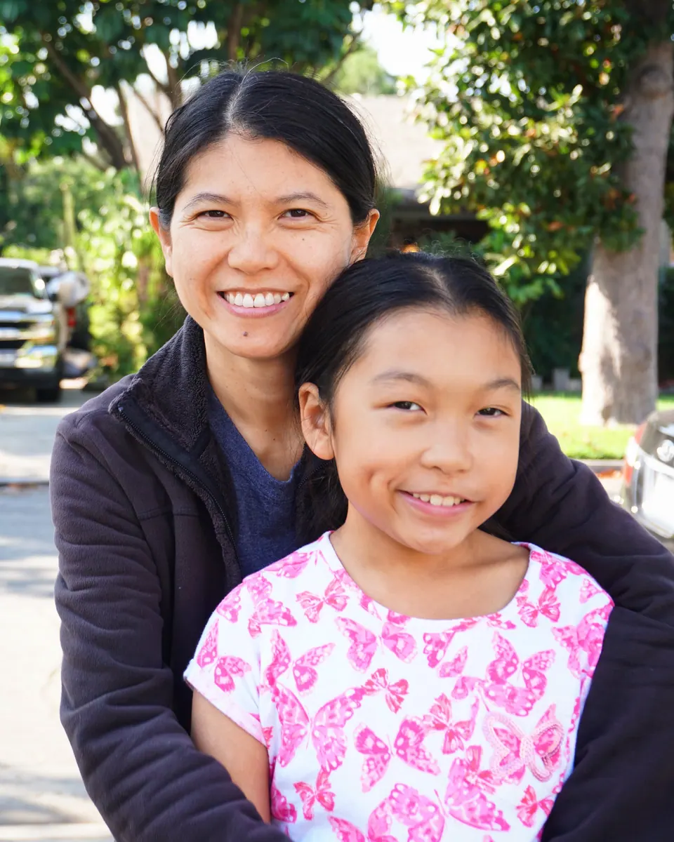 A smiling woman hugs a girl in a pink butterfly-print shirt from behind, outdoors in the sun with trees behind them.