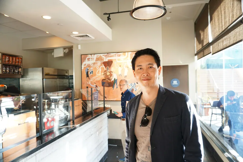 A man in a dark blazer with sunglasses hooked on his shirt stands in a bright coffee shop, a large mural of coffee roasting behind the counter.