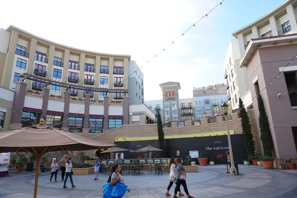 The open-air plaza of a shopping and apartment complex with umbrellas, strings of lights and a few shoppers under a pale sky.